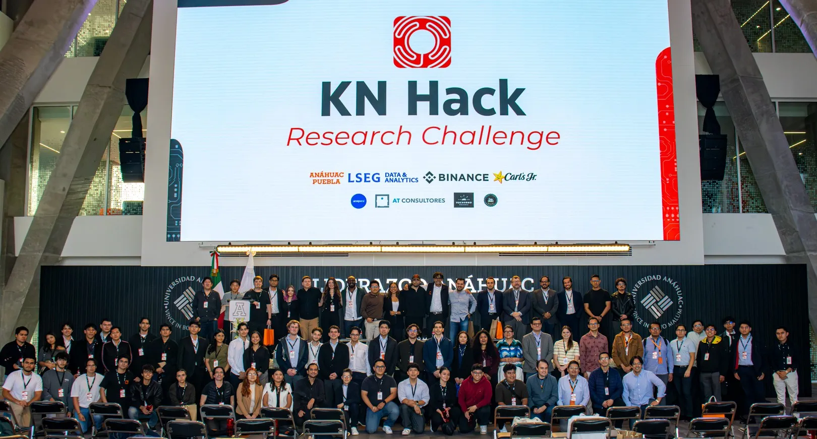 Participantes, ponentes y organizadores del Research Challenge del KN Hack 2026 en el escenario, bajo la pantalla del evento.